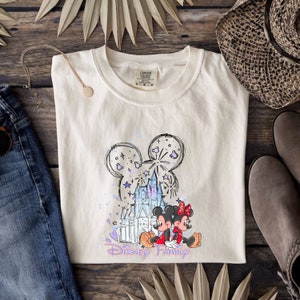 May include: A white t-shirt featuring a sketch of Mickey Mouse ears, Cinderella's castle, and Mickey and Minnie Mouse. The text "Disney Family" is printed below the image.