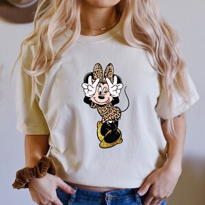 K&ouml;nnte beinhalten: Cremefarbenes T-Shirt mit einem Minnie Mouse Cartoon-Design. Minnie tr&auml;gt ein Outfit mit Leopardenmuster und zeigt mit ihren Fingern das Friedenszeichen. Ihre Schuhe sind goldfarben glitzernd. Das Design ist mittig auf der Vorderseite des Shirts.