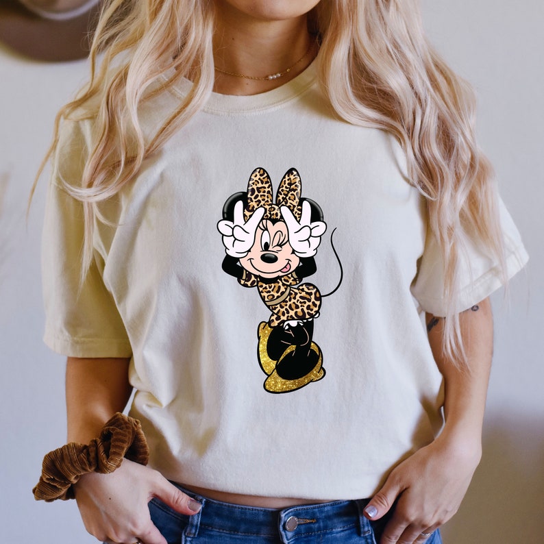 K&ouml;nnte beinhalten: Cremefarbenes T-Shirt mit einem Minnie Mouse Cartoon-Design. Minnie tr&auml;gt ein Outfit mit Leopardenmuster und zeigt mit ihren Fingern das Friedenszeichen. Ihre Schuhe sind goldfarben glitzernd. Das Design ist mittig auf der Vorderseite des Shirts.