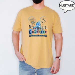 以下が含まれることがあります： マスタードイエローのTシャツに、ディズニーワールドで卒業を祝うミッキーマウスとその仲間たちのグラフィックがプリントされています。Tシャツには「Graduate from the tassel to the castle」と「2025 Senior」の文字が書かれています。