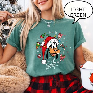 May include: Light green t-shirt featuring a cartoon Goofy character wearing a Santa hat and surrounded by Christmas decorations. The word "Goofy" is written in a script font. The speech bubble says "LIGHT GREEN".