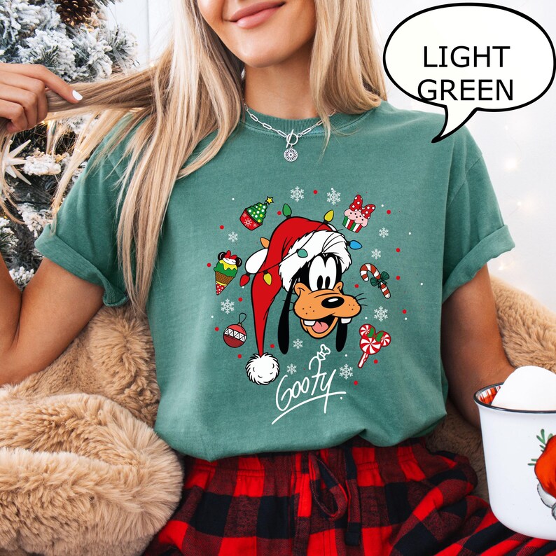 May include: Light green t-shirt featuring a cartoon Goofy character wearing a Santa hat and surrounded by Christmas decorations. The word "Goofy" is written in a script font. The speech bubble says "LIGHT GREEN".