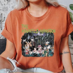 May include: Orange t-shirt with a graphic print of Mickey Mouse and friends in front of the Haunted Mansion. The text "THE HAUNTED MANSION" is printed in green above the image.