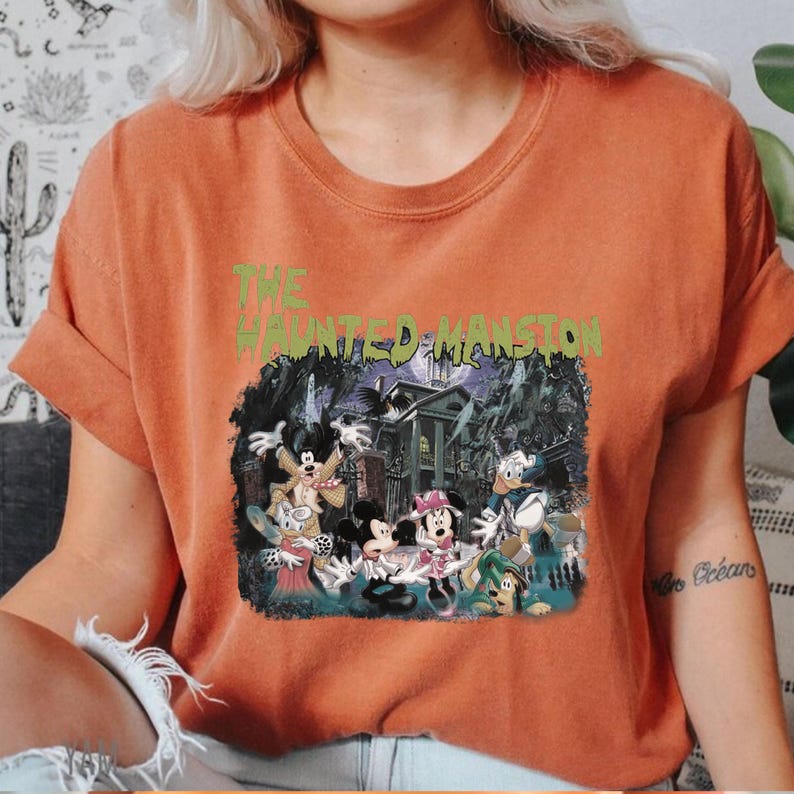 May include: Orange t-shirt with a graphic print of Mickey Mouse and friends in front of the Haunted Mansion. The text "THE HAUNTED MANSION" is printed in green above the image.