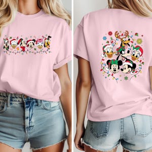 May include: A light pink t-shirt featuring a festive design with Disney characters wearing Santa hats and surrounded by Christmas lights. The front of the shirt displays a line of characters, while the back showcases a group of characters.
