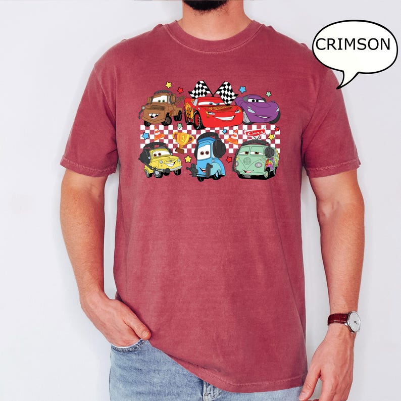 May include: A crimson red t-shirt with a graphic print of cartoon cars from the movie Cars. The cars are in a line and include Lightning McQueen, Mater, and others. The graphic is on a checkered background with stars.