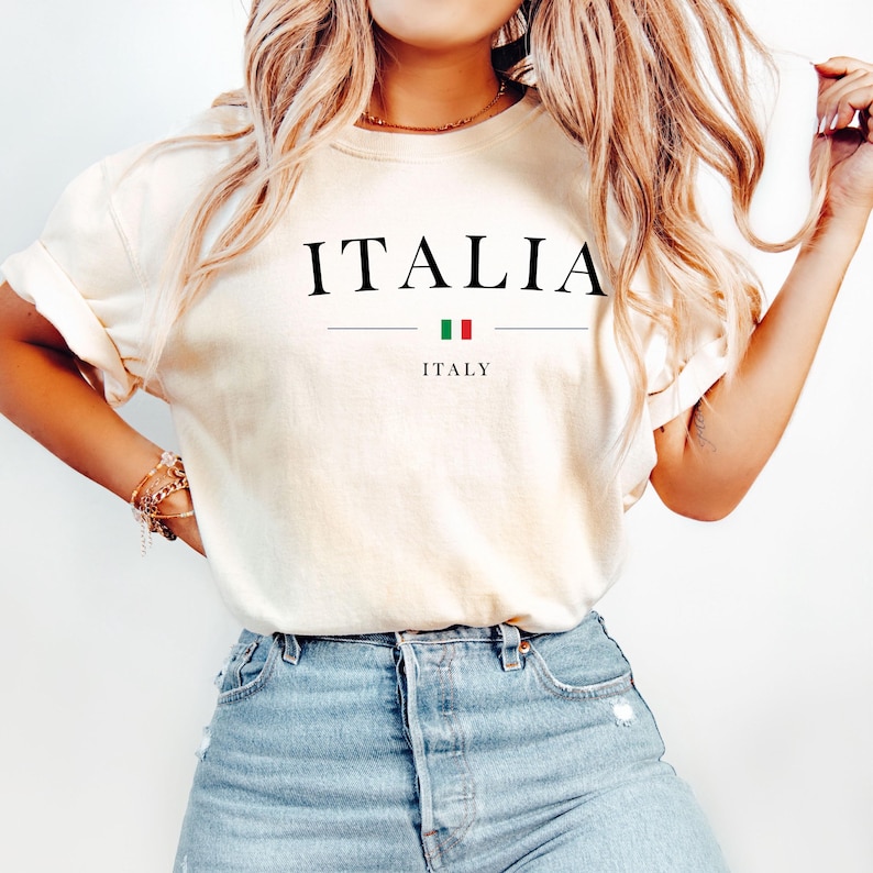May include: A white t-shirt with the word "ITALIA" printed in black letters, with the Italian flag and the word "ITALY" printed underneath.