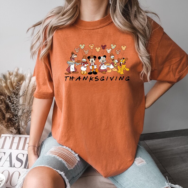 May include: Orange t-shirt with a graphic of Mickey Mouse and friends celebrating Thanksgiving. The graphic includes Mickey Mouse, Minnie Mouse, Donald Duck, Goofy, Pluto, and a turkey. The text "THANKSGIVING" is printed below the graphic.