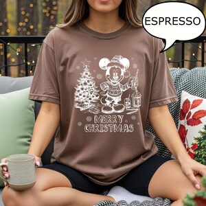 Puede incluir: Camiseta color espresso con un gr&aacute;fico blanco de Mickey Mouse con un su&eacute;ter navide&ntilde;o, sosteniendo una linterna y junto a un &aacute;rbol de Navidad. El texto "MERRY CHRISTMAS" est&aacute; debajo. La camiseta es de corte holgado.