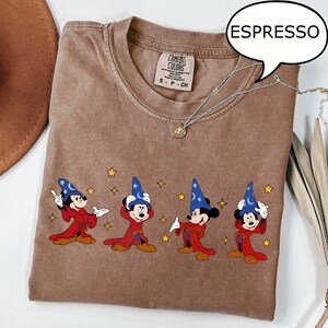 May include: Espresso-colored t-shirt featuring a graphic of Mickey Mouse in a sorcerer's outfit. The design includes Mickey in a red robe and blue wizard hat, with yellow stars. The shirt has a Comfort Colors tag.