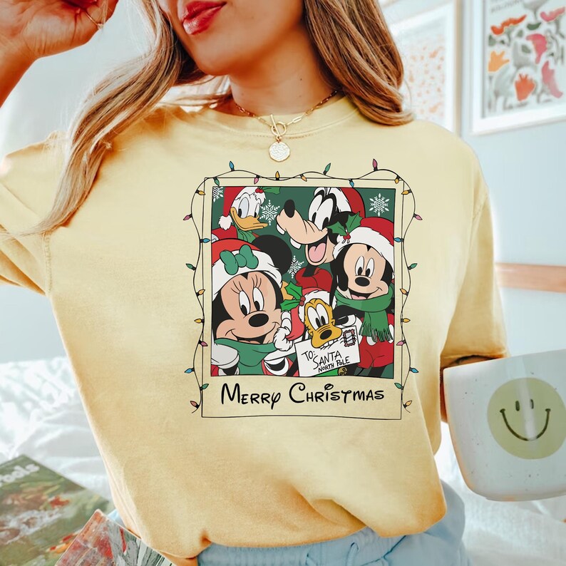 May include: A yellow t-shirt with a Christmas-themed graphic featuring Mickey Mouse, Minnie Mouse, Goofy, and Donald Duck. The graphic is a vintage-style photo with a string of Christmas lights around it. The text "Merry Christmas" is printed below the graphic.