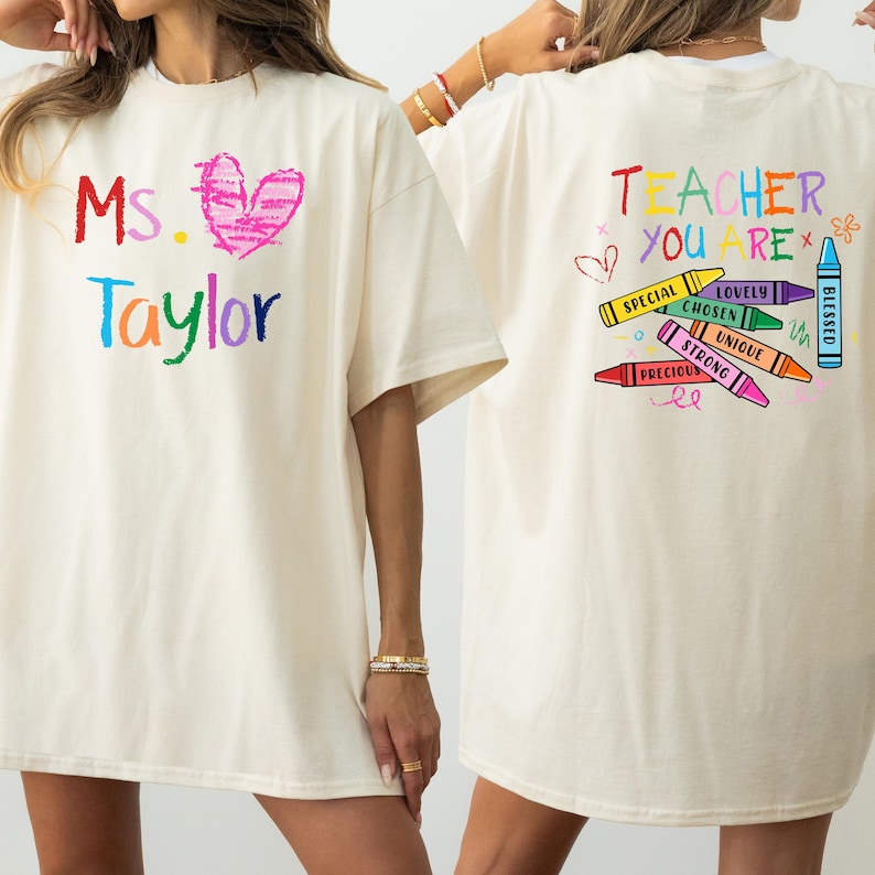 May include: Cream-colored t-shirt with colorful crayon-style text and graphics. The front features "Ms. Taylor" with a heart. The back says "Teacher You Are" with crayon-themed words like "Special" and "Blessed".