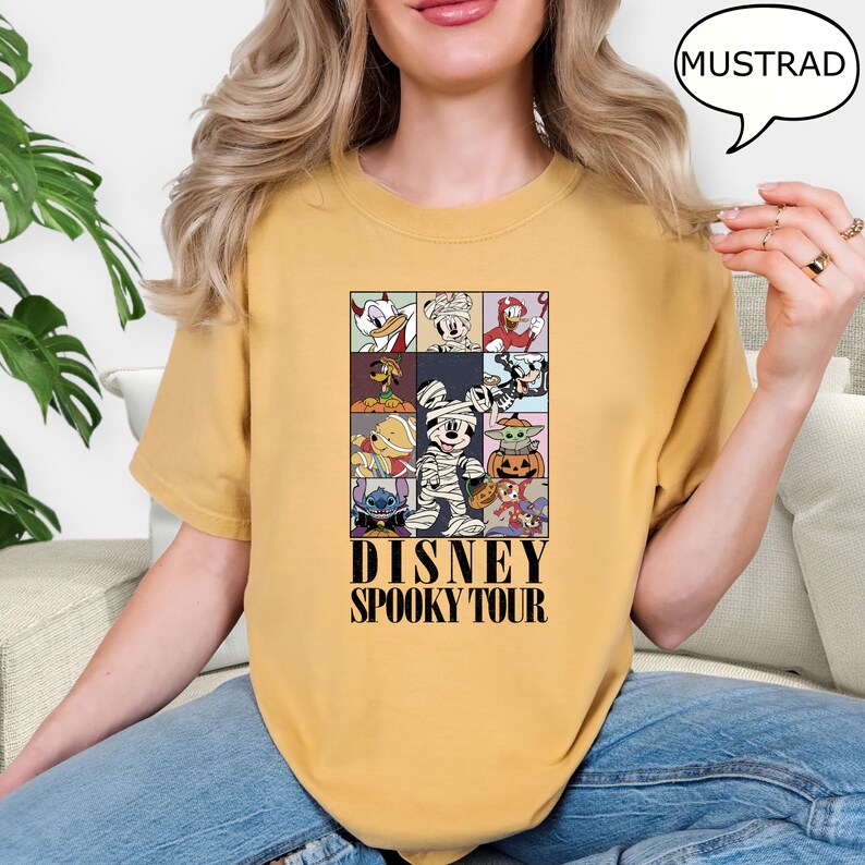 May include: Mustard-colored t-shirt featuring a grid of Disney characters in Halloween costumes. The shirt has the text "DISNEY SPOOKY TOUR" in black. The shirt is made of a soft material.