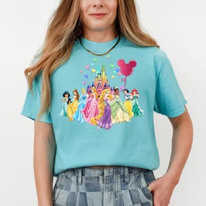 May include: A light blue t-shirt with a graphic of Disney princesses in front of Cinderella's castle. The princesses are wearing their signature dresses and there is a pink Mickey Mouse balloon in the upper right corner.