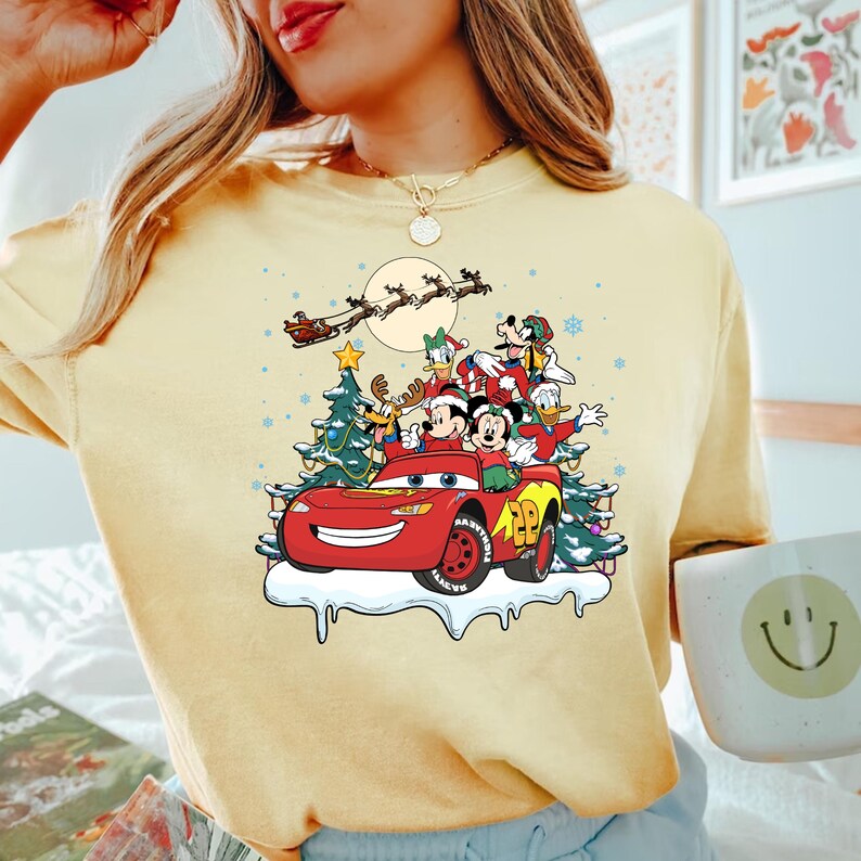 May include: A yellow t-shirt with a Christmas-themed design featuring Mickey Mouse, Donald Duck, Goofy, and Pluto riding in a red Lightning McQueen car. The design also includes a Christmas tree, snow, and a moon with Santa Claus flying in a sleigh.