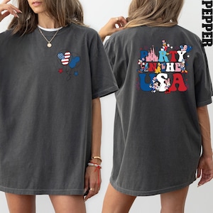 K&ouml;nnte beinhalten: Dunkelgraues T-Shirt mit einer Grafik von Ballons in Rot, Wei&szlig; und Blau und dem Text "Party in the USA" mit Zeichentrickfiguren. Das Shirt hat eine lockere Passform und kurze &Auml;rmel.