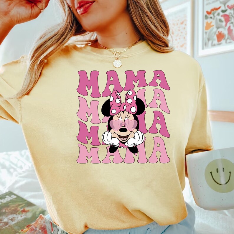 Puede incluir: Una camiseta amarilla con un gr&aacute;fico rosa de Minnie Mouse con gafas de sol en forma de coraz&oacute;n y la palabra "MAMA" repetida cuatro veces en una fuente retro.