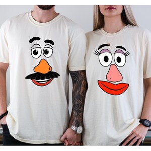 May include: Two cream-colored t-shirts featuring cartoon faces. One shirt has a black mustache, orange nose, and a red mouth. The other shirt has eyelashes, a pink nose, and a red mouth. The shirts are a matching set.