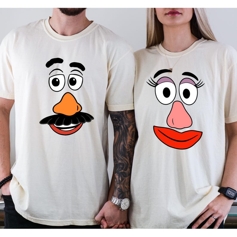 May include: Two cream-colored t-shirts featuring cartoon faces. One shirt has a black mustache, orange nose, and a red mouth. The other shirt has eyelashes, a pink nose, and a red mouth. The shirts are a matching set.