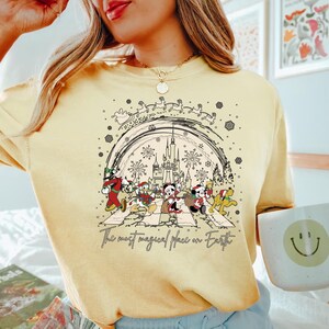 Puede incluir: Una camiseta amarilla con una ilustraci&oacute;n en blanco y negro de Mickey Mouse y sus amigos caminando por una calle con un castillo en el fondo. El texto "The most magical place on Earth" est&aacute; impreso debajo de la ilustraci&oacute;n.