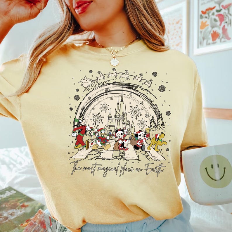 Puede incluir: Una camiseta amarilla con una ilustraci&oacute;n en blanco y negro de Mickey Mouse y sus amigos caminando por una calle con un castillo en el fondo. El texto "The most magical place on Earth" est&aacute; impreso debajo de la ilustraci&oacute;n.