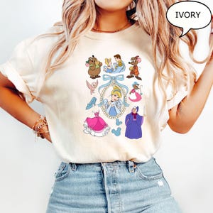 Puede incluir: Camiseta de color marfil con un gr&aacute;fico colorido de Cenicienta y otros personajes de la pel&iacute;cula animada. El dise&ntilde;o incluye a Cenicienta con un vestido azul, un vestido rosa y otros elementos ic&oacute;nicos. La palabra "IVORY" est&aacute; en una burbuja.