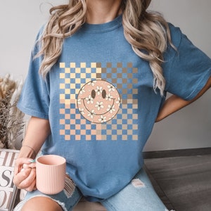 Peut inclure: Un t-shirt bleu avec un motif &agrave; carreaux en jaune, marron et p&ecirc;che. Un smiley rose avec des fleurs blanches est centr&eacute; sur le motif &agrave; carreaux.