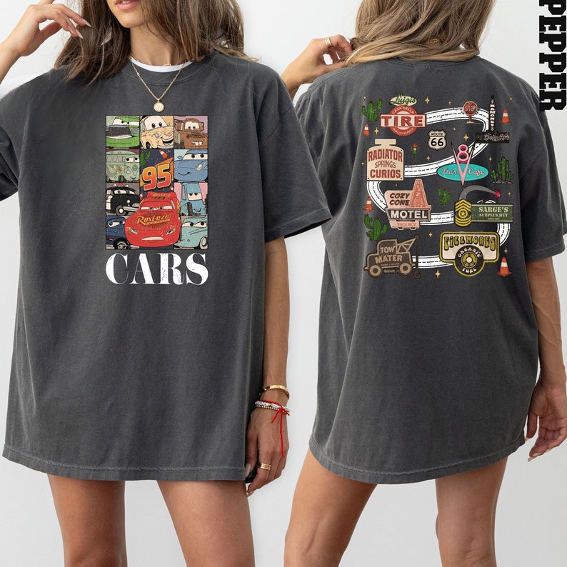 Puede incluir: Una camiseta de gran tama&ntilde;o de color gris oscuro con un gr&aacute;fico tem&aacute;tico de Cars en la parte delantera y un gr&aacute;fico tem&aacute;tico de la Ruta 66 en la parte posterior. El gr&aacute;fico delantero presenta un collage de personajes de Cars, incluidos Rayo McQueen, Mate y otros. El gr&aacute;fico trasero presenta un mapa de Radiador Springs con varios puntos de referencia y negocios.
