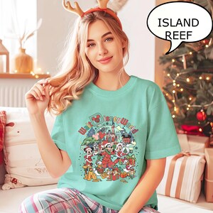 May include: A light teal t-shirt featuring a colorful graphic of Disney characters in Christmas attire, with the text "Walt Disney World". The shirt also has the text "Island Reef" in a speech bubble. The shirt is worn by a person.
