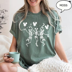 May include: Moss green t-shirt featuring a white graphic of three dancing skeletons holding Mickey Mouse-shaped balloons. The shirt has rolled-up sleeves and a relaxed fit. The word "MOSS" is in a speech bubble.