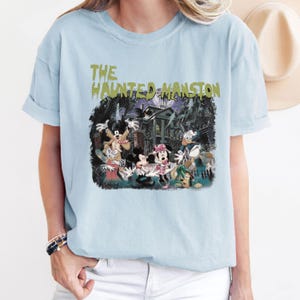May include: Light blue t-shirt with a graphic print of Mickey Mouse and friends in front of the Haunted Mansion. The text "The Haunted Mansion" is printed in green on the shirt.