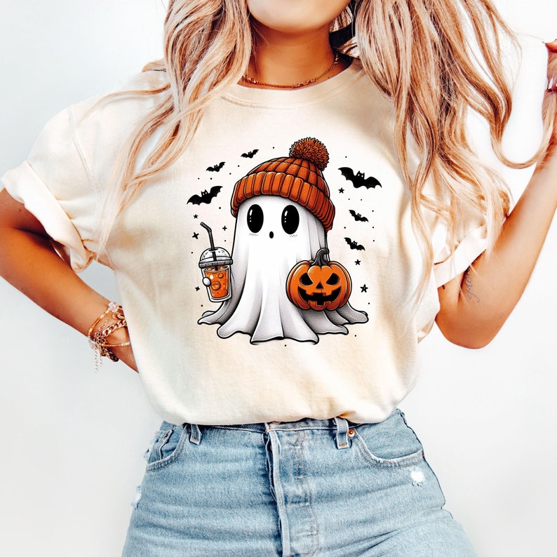 May include: A white t-shirt featuring a cute cartoon ghost wearing an orange and brown knitted hat, holding a pumpkin and a cup of orange drink. The ghost is surrounded by bats and stars.
