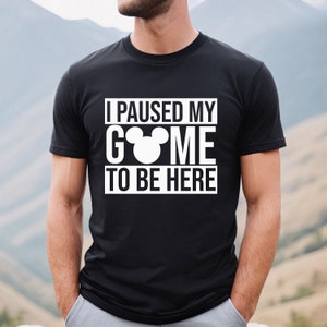 May include: Black t-shirt with the text "I PAUSED MY GAME TO BE HERE" in white, with a white Mickey Mouse head replacing the "O" in "GAME". The shirt is a crew neck and has short sleeves.