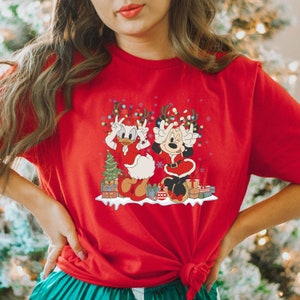 Puede incluir: Camiseta roja con un gr&aacute;fico navide&ntilde;o con Mickey Mouse y Daisy Duck con gorros de Pap&aacute; Noel y sosteniendo luces navide&ntilde;as. El gr&aacute;fico tambi&eacute;n incluye un peque&ntilde;o &aacute;rbol de Navidad y regalos.