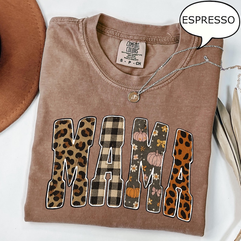 May include: Espresso-colored t-shirt with the word "MAMA" in a decorative font. The letters have a leopard print, plaid, and floral pattern. A silver necklace with a pendant is on the shirt.