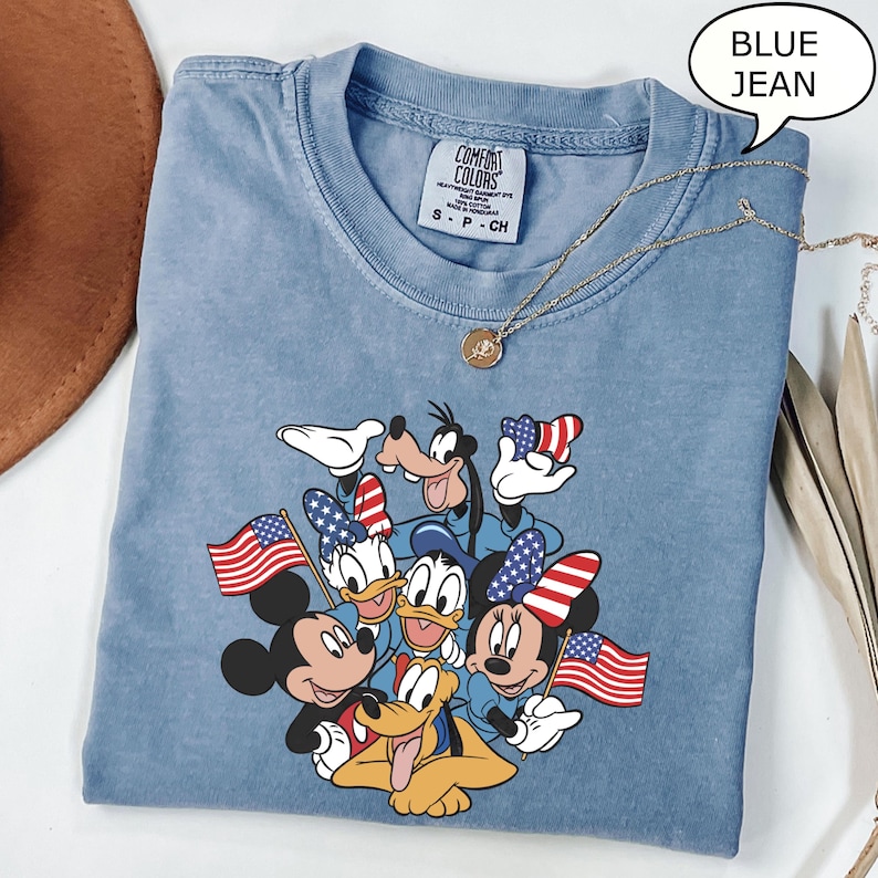 May include: A light blue Comfort Colors t-shirt featuring a graphic of Mickey Mouse, Minnie Mouse, Donald Duck, Daisy Duck, Goofy, and Pluto, all holding American flags and wearing patriotic accessories. A gold necklace and the text "BLUE JEAN" are also visible.