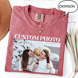 May include: Crimson-colored t-shirt with a photo print and the text "CUSTOM PHOTO." The shirt features a crew neck and short sleeves. A silver necklace with a pendant is draped across the shirt. The shirt is a Comfort Colors brand.