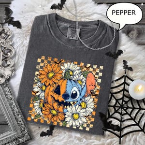 May include: Dark gray t-shirt with a Halloween-themed graphic. The design features a pumpkin and a blue character surrounded by orange and white flowers. The shirt has a crew neck and short sleeves. The word "PEPPER" is in a speech bubble.