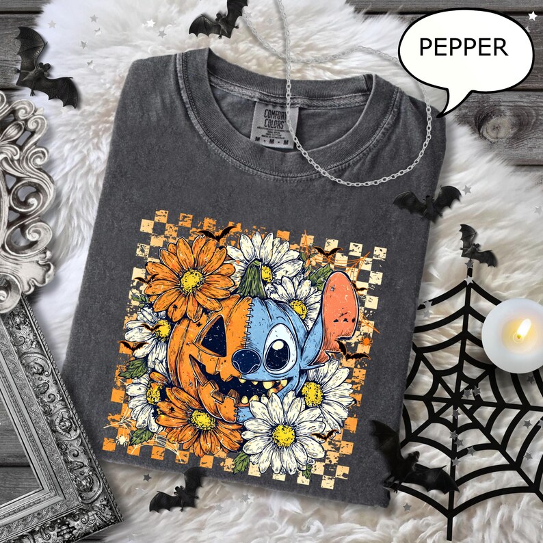 May include: Dark gray t-shirt with a Halloween-themed graphic. The design features a pumpkin and a blue character surrounded by orange and white flowers. The shirt has a crew neck and short sleeves. The word "PEPPER" is in a speech bubble.