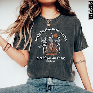 Op de afbeelding: Een donkergrijs T-shirt met een afbeelding van drie skeletten die rond een kampvuur dansen. De tekst "they're burning all the witches" staat boven de afbeelding en "even if you aren't one" eronder. Het woord "reputation" staat in een kleiner lettertype onder de tekst.