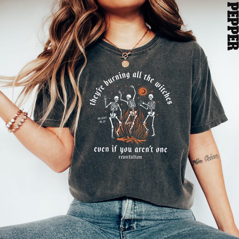 Op de afbeelding: Een donkergrijs T-shirt met een afbeelding van drie skeletten die rond een kampvuur dansen. De tekst "they're burning all the witches" staat boven de afbeelding en "even if you aren't one" eronder. Het woord "reputation" staat in een kleiner lettertype onder de tekst.