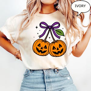 May include: Ivory-colored t-shirt featuring a Halloween-themed graphic. The design showcases two orange jack-o'-lanterns with smiling faces, connected by stems and a purple bow. A green leaf and stars complete the festive illustration.