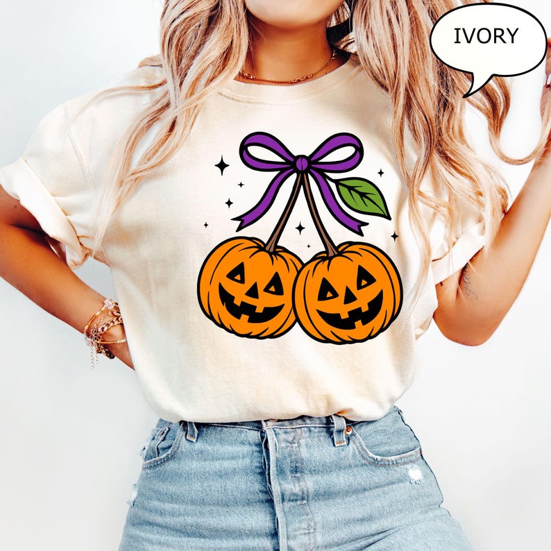 May include: Ivory-colored t-shirt featuring a Halloween-themed graphic. The design showcases two orange jack-o'-lanterns with smiling faces, connected by stems and a purple bow. A green leaf and stars complete the festive illustration.