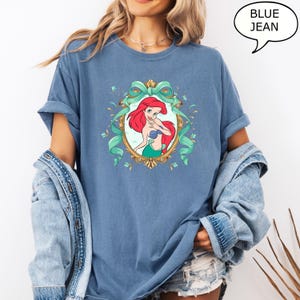 May include: Blue t-shirt featuring a cartoon illustration of a mermaid with red hair, a blue top, and a green tail, framed by a gold oval and teal ribbons. The text "BLUE JEAN" is in a speech bubble.