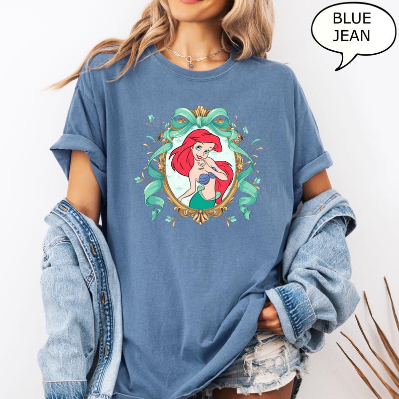 May include: Blue t-shirt featuring a cartoon illustration of a mermaid with red hair, a blue top, and a green tail, framed by a gold oval and teal ribbons. The text "BLUE JEAN" is in a speech bubble.