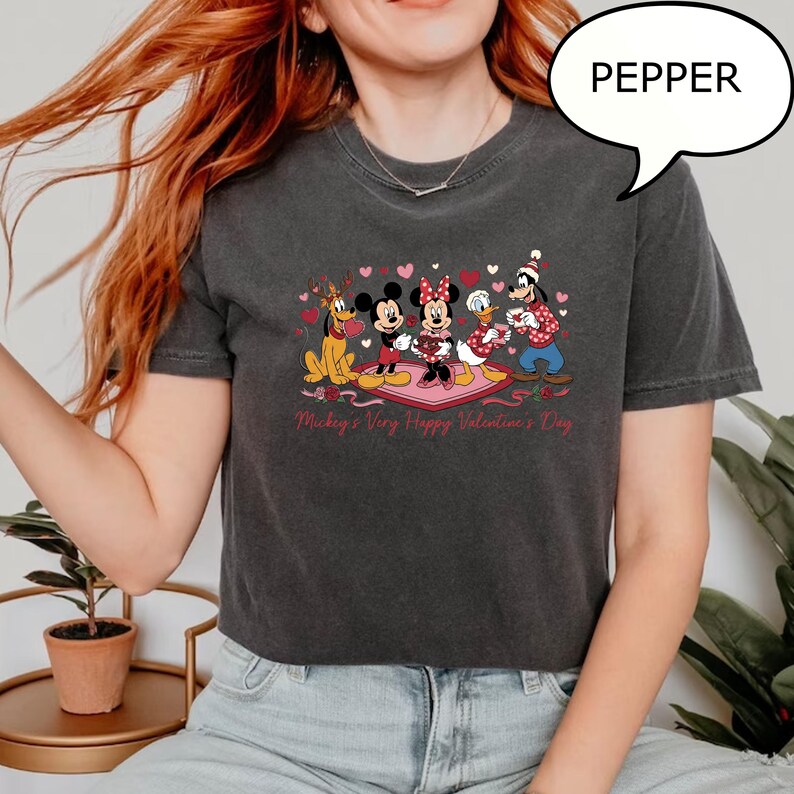 Puede incluir: Camiseta gris oscuro con un dise&ntilde;o de San Valent&iacute;n con Mickey Mouse, Minnie Mouse, el Pato Donald, Goofy y Pluto. El texto "Mickey's Very Happy Valentine's Day" est&aacute; impreso debajo del dise&ntilde;o. Un bocadillo con la palabra "PEPPER" est&aacute; en la esquina superior derecha.