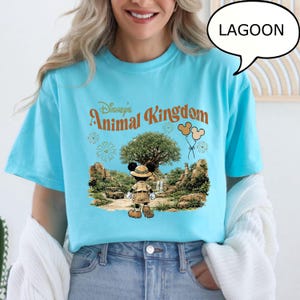 May include: A light blue t-shirt featuring the text "Disney's Animal Kingdom" in orange and a graphic of Mickey Mouse in safari attire. The graphic includes a tree, balloons, and fireworks.