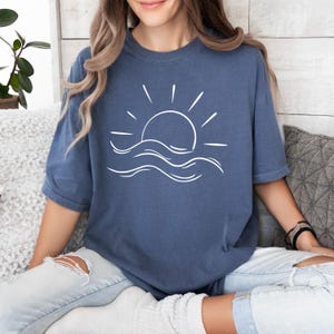 May include: A blue oversized t-shirt with a white graphic of a sun setting over waves.