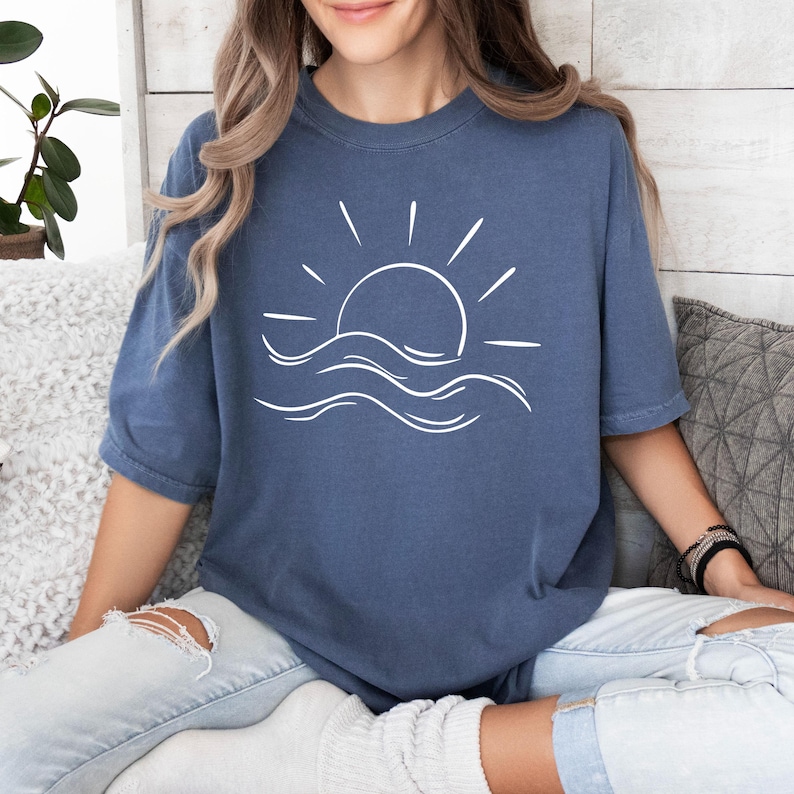 May include: A blue oversized t-shirt with a white graphic of a sun setting over waves.