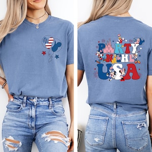 K&ouml;nnte beinhalten: Hellblaues T-Shirt mit patriotischem Design. Auf der Vorderseite ist ein Mickey Mouse-f&ouml;rmiger Ballon mit amerikanischem Flaggenmuster zu sehen, und auf der R&uuml;ckseite steht "Party in the USA" mit Mickey Mouse-Figuren und Feuerwerk.